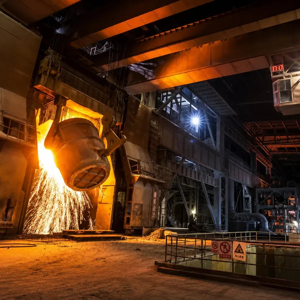 Steel ladle pouring molten steel inside a large industrial steel plant, with intense sparks, overhead crane systems, glowing furnace light, and heavy-duty factory structures in a high-temperature steelmaking environment.